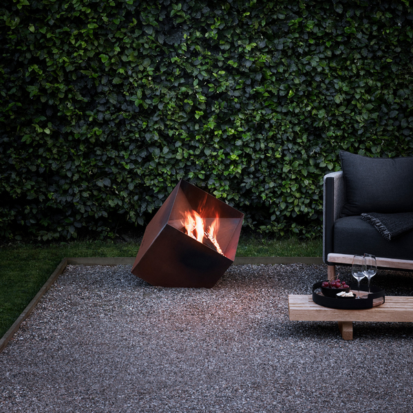 Firecube Fire Pit by Eva Solo with a fire burning in an outdoor setting on a gravel surface next to a lawn and hedged area. A grey chair next to it and a couple of almost empty wine glasses on a low bench table with a bowl of red grapes and crackers.