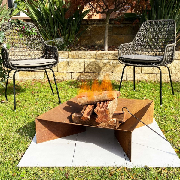Fuji Rust FirePit and poker with a fire burning in a garden setting on lawn with 2 chairs and plants in the background.