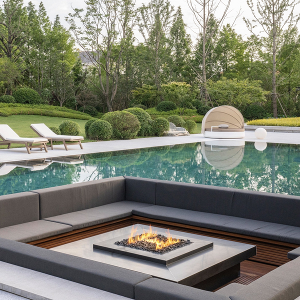 Galio Gas Fire Pit H Insert - 60cm Square next to a swimming pool with stainless steel table surround, wooden bench seating all around it, pool side sunloungers on other side of the pool with a manicured lawn, round sculptured shrubs and trees in the backdrop.