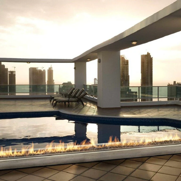 Galio Gas Fire Pit Insert Linear Automatic with fire burning at a rooftop poolside, tiles surrounding pool, sun lounges set up and glass fencing. Highrise buildings in the background and a view of the ocean can be seen in the not too far distance.