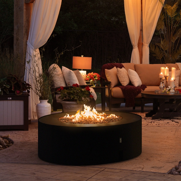 Galio Star Black Automatic Outdoor Gas Fireplace in a patio setting on brick pavers, set in front of a formal style lounge with plenty of cushions, a coffee table set with a silver tray and 5 pillar candles, a lamp and plants around.