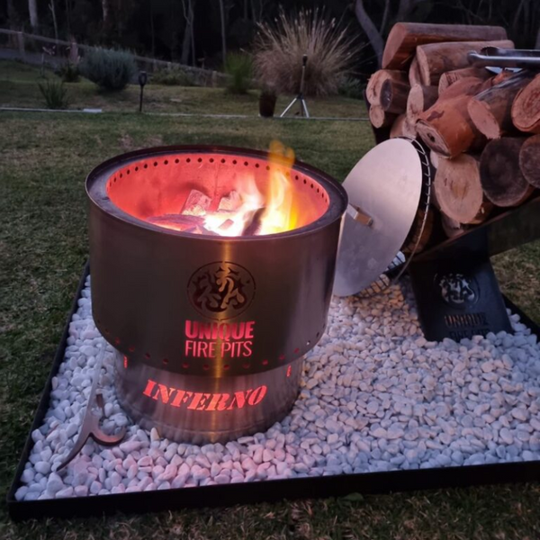 A stainless steel Inferno smokeless fire pit shown with a fire burning inside it, placed on a bed of stones outdoors next to a stack of wood.