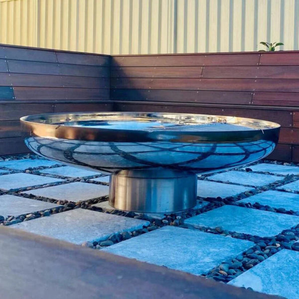 Cauldron round shaped stainless-steel fire pit on cement tiles with small pebbles and a wooden seating area in a backyard setting.