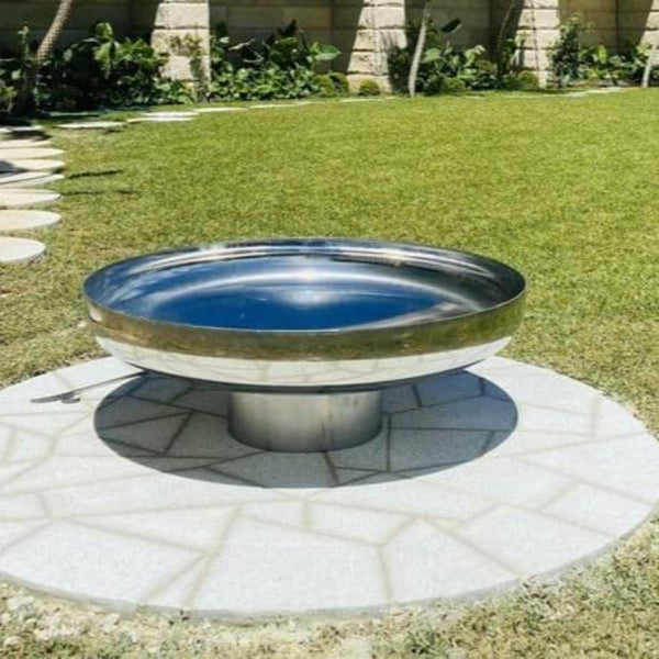 A polished mirror finish stainless steel fire pit with a circular shape, situated on a paved area with grass and plants in the background.