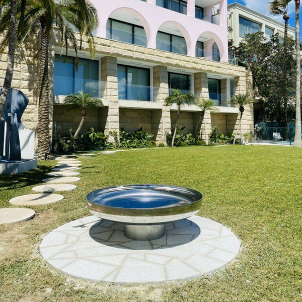A polished stainless steel fire pit placed on a grassy area with a building and palm trees in the background.