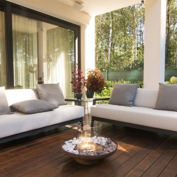 Tondo Commerce Ethanol Fire Pit with fire burning in an outdoor area on a wood deck, with lounges and cushions, couple of pot plants and garden view with ferns and trees in the background.