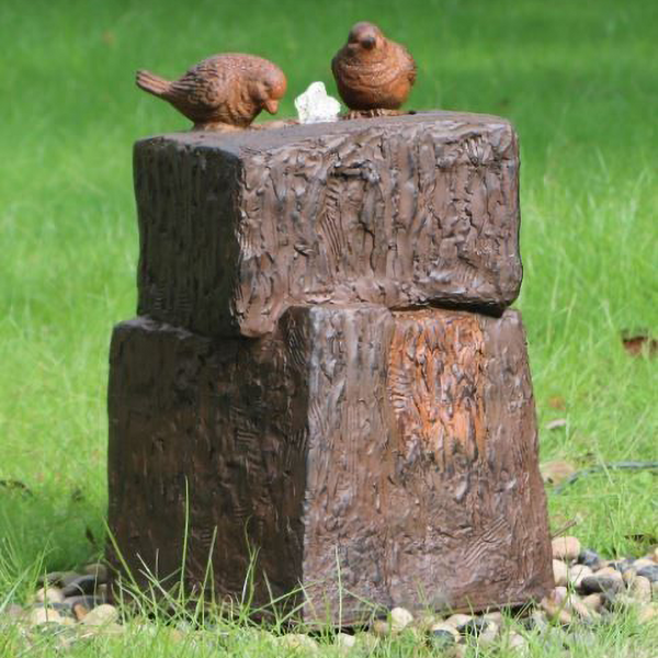 Two Dicky Birds sitting in a rock pool Water Fountain in a garden setting with pebbles, and green grass with dandelions surrounding it.
