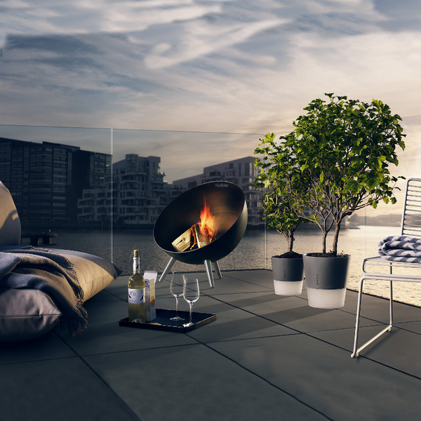 Eva Solo Fireglobe Fire Pit with fire burning on a large, tiled floor patio with glass fencing, couple of pot plants, a large beanbag type chair, a tray with bottle of wine, 2 glasses and a box of crackers. A body of water with hig rise apartment buildings in the background.