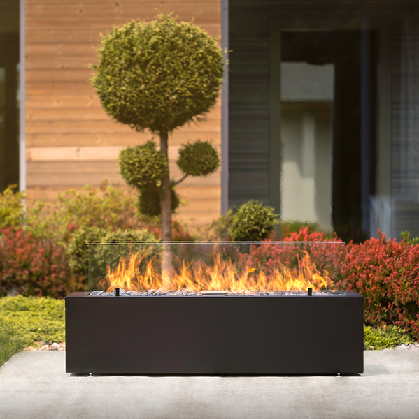 Galio Linear Automatic Black Gas Fire Pit on a cement surface set up with a fire burning surrounded with a ground cover, low shrubs and topiary trees in front of a house.
