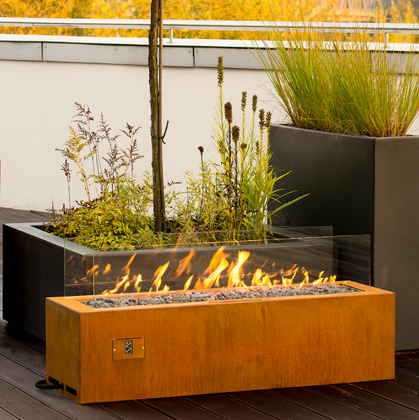Galio Linear Automatic Corten coloured Gas Fire Pit with small flames burning on a wooden deck area in front of a couple of planters with tree and plants in.