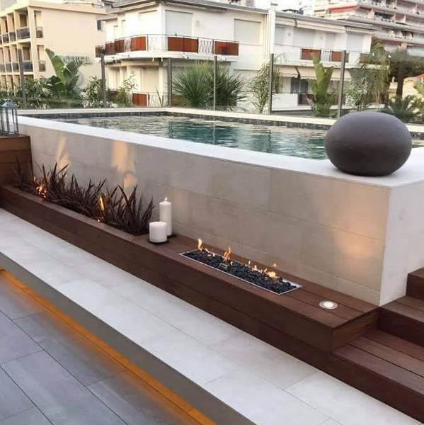Galio Gas Fire Pit Insert Linear Automatic in a hotel rooftop beside a swimming pool with decking and cement tiles surrounding it, glass safety fencing, candles, lamp and small plants around and other motels in the background, treetops can be seen between them.