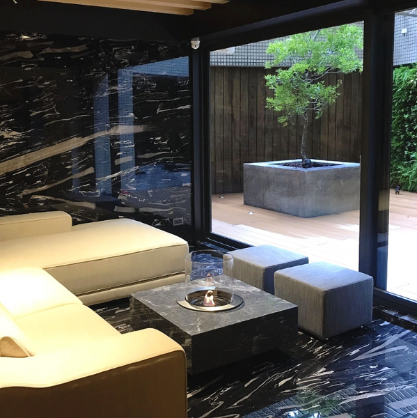 Rondo Commerce Ethanol Burner Australia at a private home in a sunroom with lounge suite and ottomans around it, room leads to a deck are where a tree can be seen in a large planter and vines hanging over a fence.