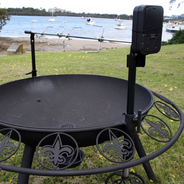 Spit Rotisserie Support Poles set up for cooking on the Ultimate BBQ Fire Pit. On a lawn area beside a body of water with boats.