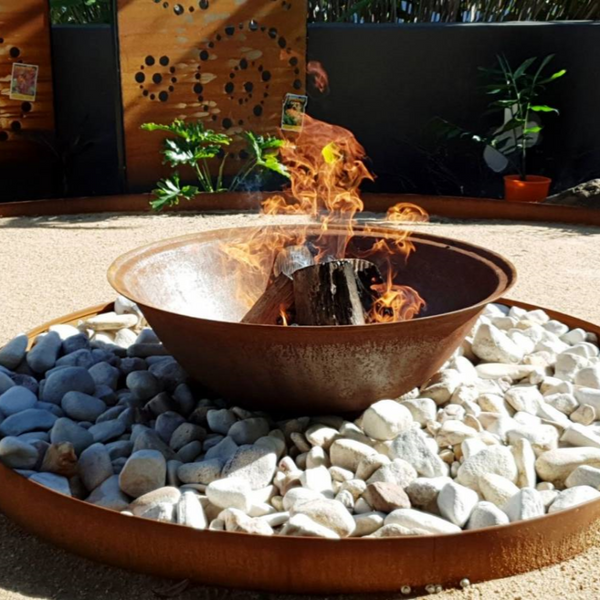 A Rust colour cast iron round basin fire pit on stand with a fire burning in a rock surround and in a garden setting.