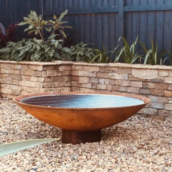 A round, rust-colored cast iron fire pit, placed on a bed of pebbles, set against a garden backdrop. The fire pit has a smooth, solid appearance with a slightly elevated rim.