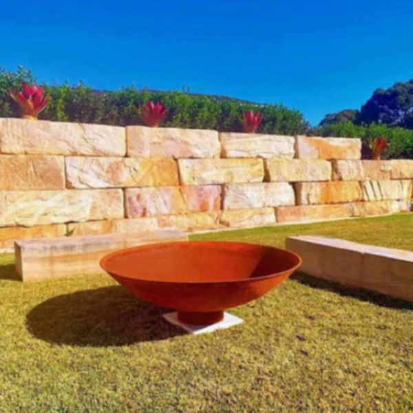 The Cauldron Fire Pit 1500mm in garden setting with large stone bench seating, on a cement tile and grass area, plants and blue sky in the background.
