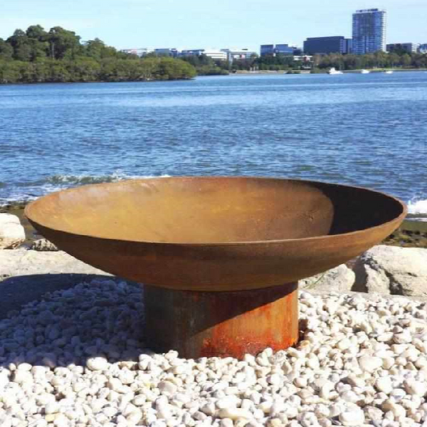 The Rust coloured Cauldron Cast iron Fire Pit with a gravel and rocky surrounding, beside a body of water with buildings and trees in the background.