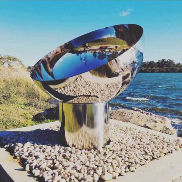 The Goblet Stainless Steel Fire Pit beside a body of water with trees in background