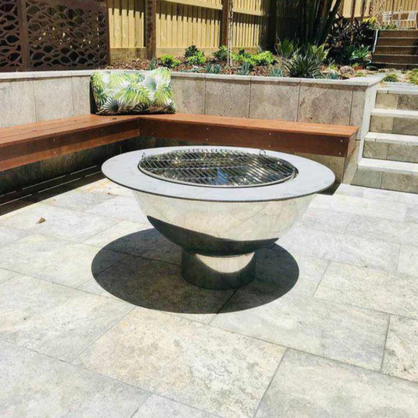 The Teppanyaki Stainless Steel Fire Pit and grill in a private garden setting with cement tiled area and wooden bench seating and plants in background.