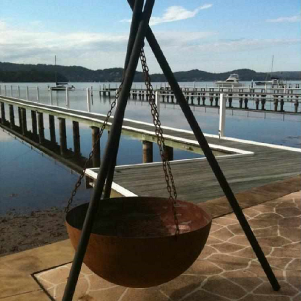 The Tripod Fire Pit with Goblet bowl on a paved area beside the sea, with several jetties, trees and boats in the background.