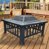 Square Metal Fire Pit Outdoor Heater set in a table with a mesh screen top with a faux-stone finish and set on a wood decking with matching wooden bench, planter and fencing, there's a house, garden, jetty and body of water in the backdrop.