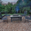 A Square Metal Fire Pit set in a table with a mesh screen top with a faux-stone finish and set on a pebbled surface with 4 metal chairs inside wooden sleeper edging around the firepit, grass and trees in the backdrop.
