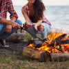 Two people crouching by a campfire outdoors near the water, using a long-handled black metal tool to tend the flames. One person wears a red plaid shirt and jeans, and the other wears a matching red plaid shirt with ripped jeans.