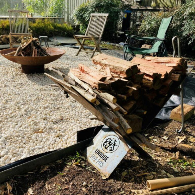 Fire pit bowl with wood stacked on a log rack with 'Unique Fire Pits' logo branding in a garden setting