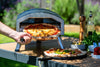 Person taking a pizza out of an outdoor pizza oven with another pizza on a stone tray nearby.