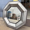 Viking Octagonal shaped Metal Mirror on cement floor and leaning on a wall in a warehouse with a few piles of stacked wooden pallets showing in the reflection of the mirror.