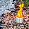 A portable stainless steel fire pit with fire burning on river rocks beside a river or creek body of water.