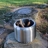 A cylindrical stainless steel fire pit with perforations for ventilation with kindling wood inside ready to burn set on a large rock, the grill on another rock next to it with rocky and grassy surrounds. 