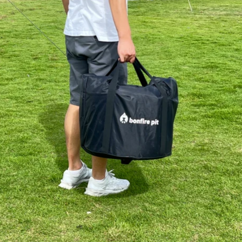 Someone carrying a large black carry bag with the word's bonfire pit and a logo on it, on a green grass area.