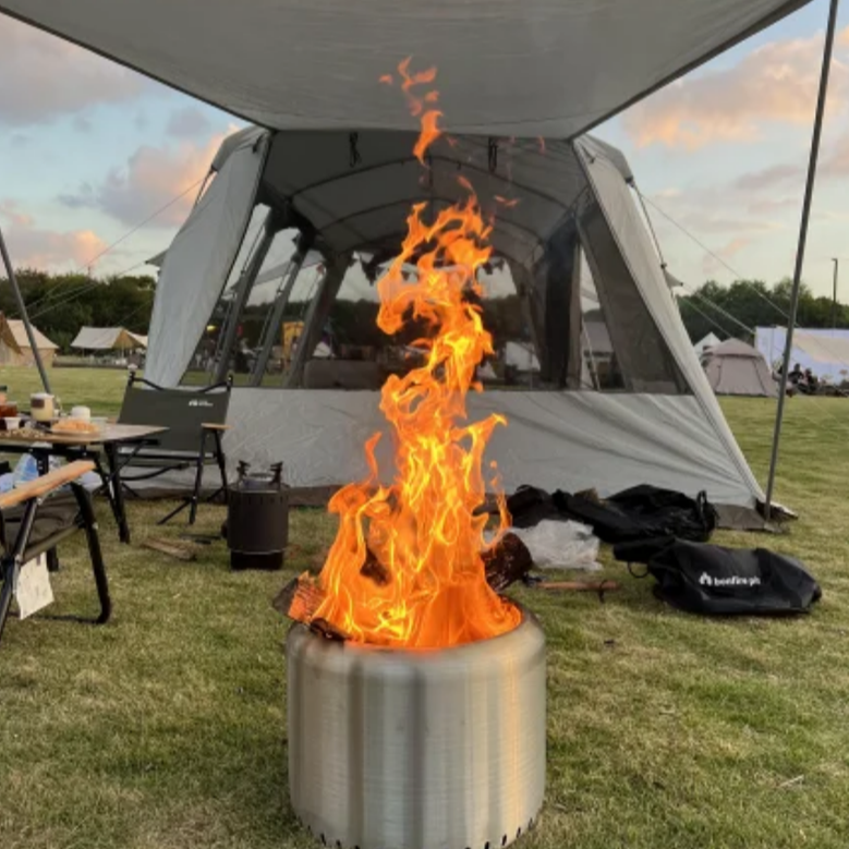 A cylindrical stainless steel fire pit with fire burning on a grass area in front of a campsite with tent and picnic table and chairs and more tents set up in the background.