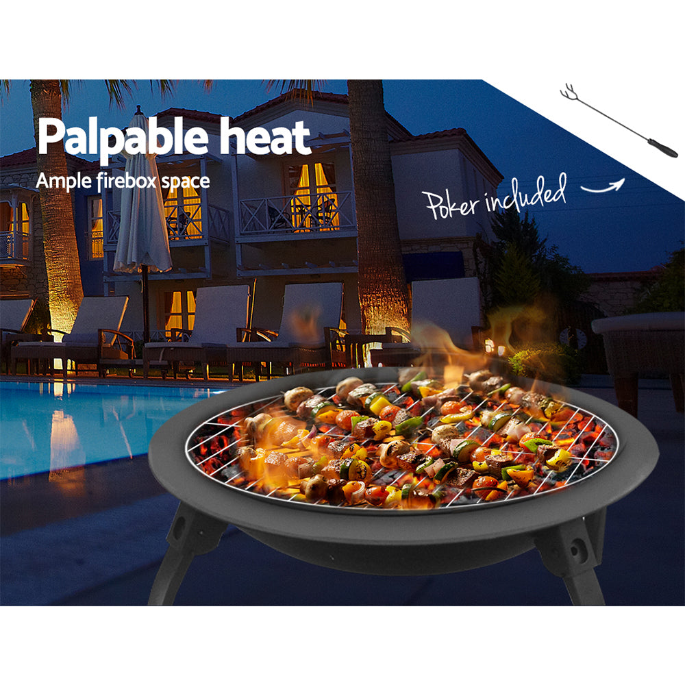 A portable black fire pit with a foldable design, cooking meat and vegetable kebabs, set next to a poolside with chairs around and a house in the background.