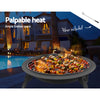 A portable black fire pit with a foldable design, cooking meat and vegetable kebabs, set next to a poolside with chairs around and a house in the background.