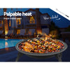 A portable black fire pit with a foldable design, cooking meat and vegetable kebabs, set next to a poolside with chairs around and a house in the background.