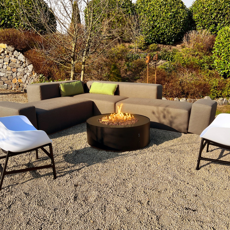 Galio Star Black Automatic Outdoor Gas Fireplace in an outdoor setting on a gravel surface with a sofa lounge and couple of chairs, garden with rock wall and lots of trees and a couple of bird feeders in the background.