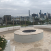 A round, star-shaped outdoor gas fireplace insert with slate flooring and cement rounded seating around it on a roof top area with glass panel fencing. A city with lots of buildings and a football field can be seen in the view behind.