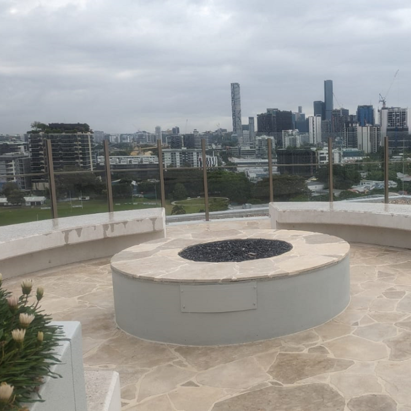 A round, star-shaped outdoor gas fireplace insert with slate flooring and cement rounded seating around it on a roof top area with glass panel fencing. A city with lots of buildings and a football field can be seen in the view behind.