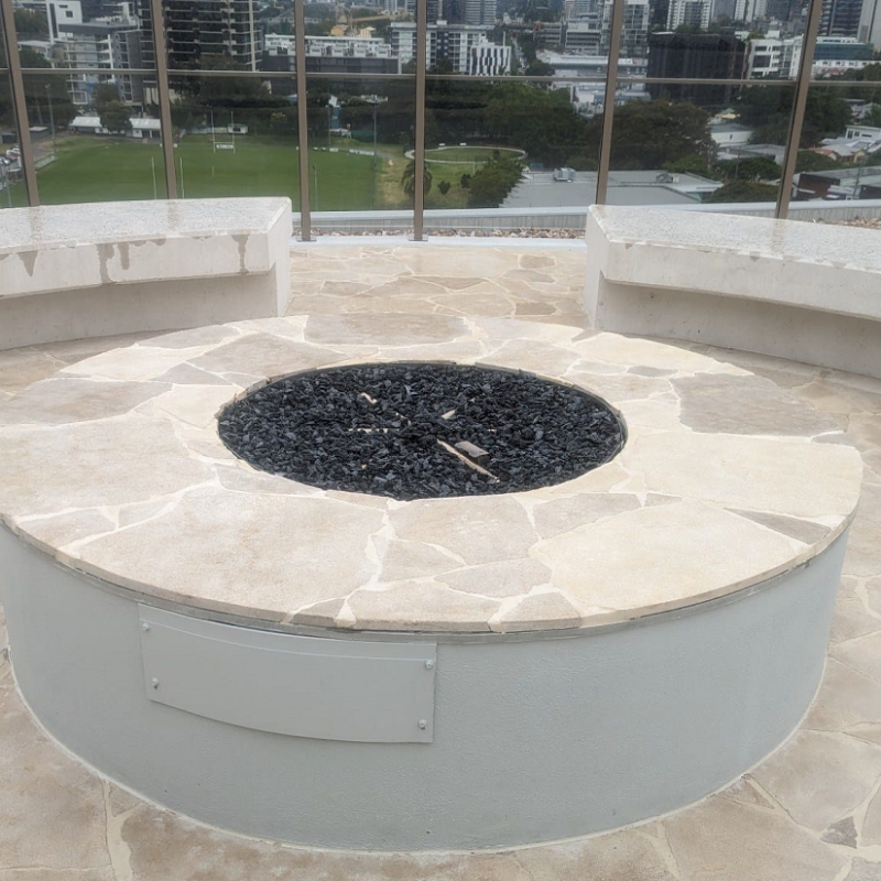 Close up view of a round, star-shaped outdoor gas fireplace insert with slate flooring and fire pit surround, with cement seating on a roof top area with glass panel fencing. A city with buildings and football field can be seen in the backdrop.