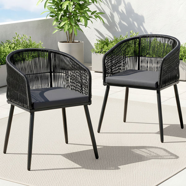 Two black wicker chairs with dark cushions on a patio with plants in the background.