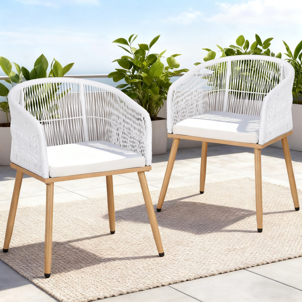 Two white wicker chairs with wooden legs on a patio with plants in the background.