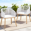 Two white wicker chairs with wooden legs on a patio with plants in the background.
