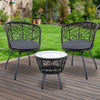 A three-piece outdoor bistro set comprising two high-backed chairs with black cushions and a round rattan table with a clear glass top on a black frame, on a wooden deck in a garden setting with green lawn, trees and flowers in the background.