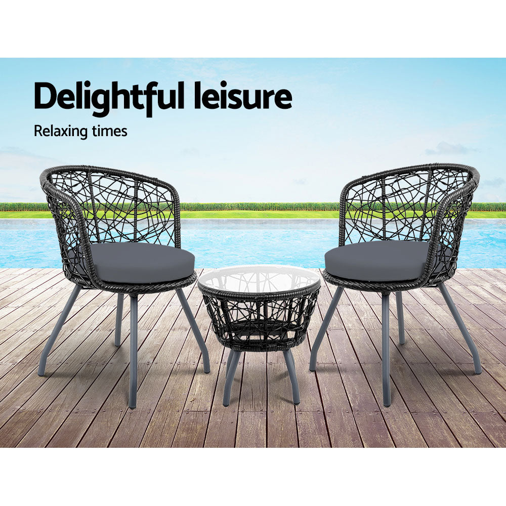 A three-piece outdoor bistro set consisting of two chairs with black cushions and a round rattan table with a clear glass top on a black frame, set on a wooden deck area in front of a poolside with grass, hedging and blue sky in the backdrop.