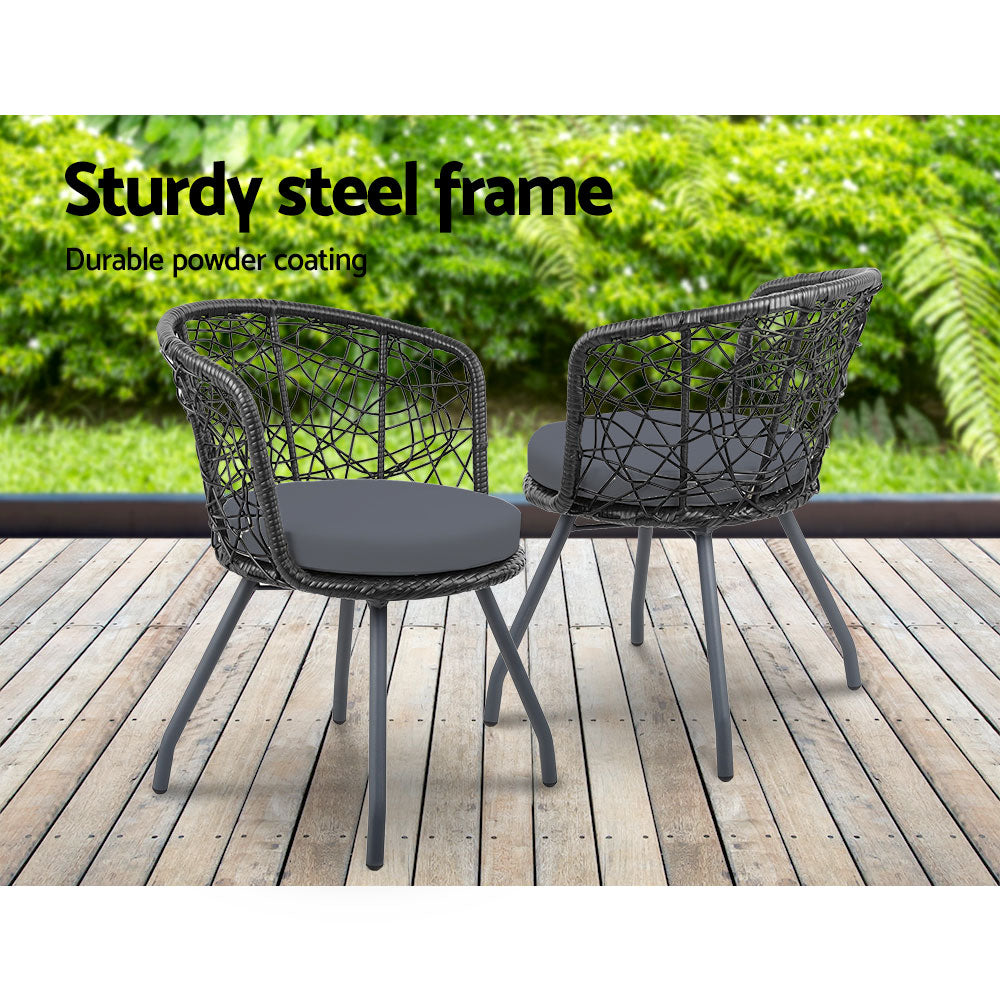 Two black rattan chairs with black cushions on a black frame, set on a wooden decking in front of a grass and greenery in the backdrop.