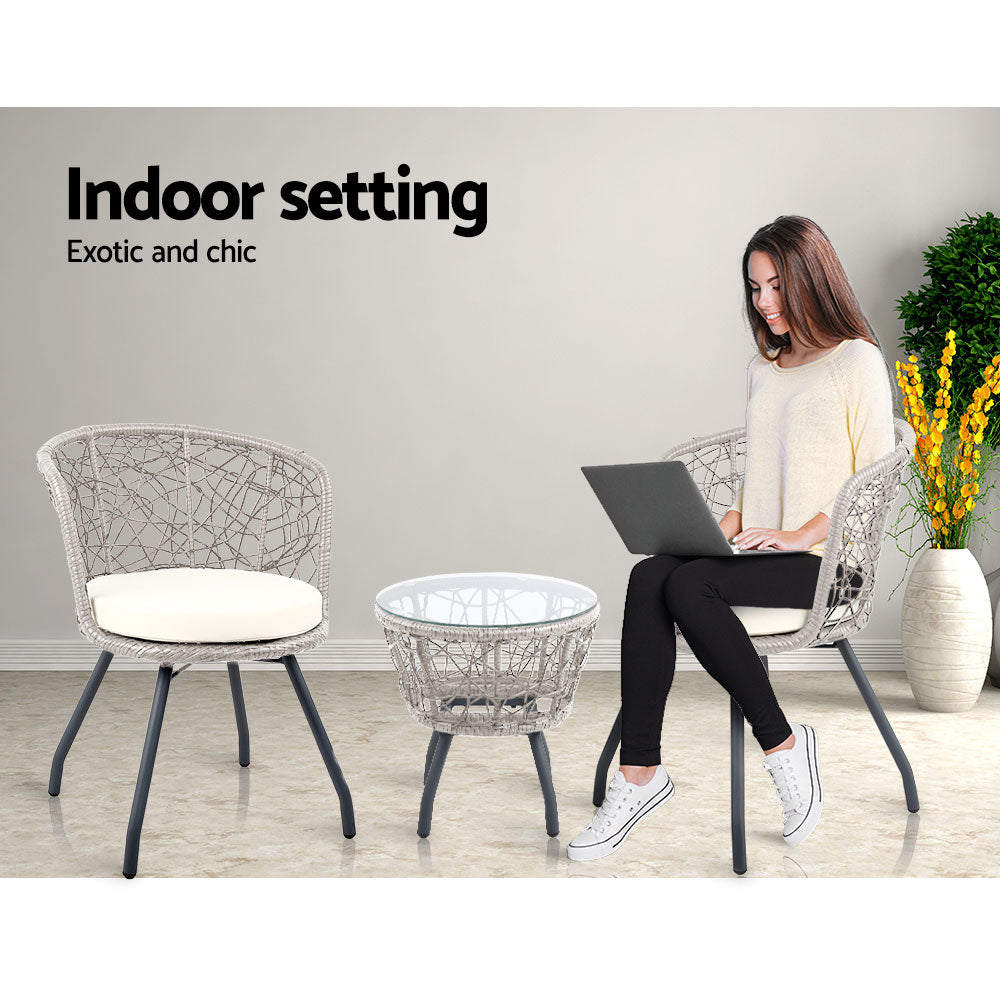 A three-piece bistro set consisting of two chairs with beige cushions and a round rattan table with a clear glass top on a grey frame, set on a marble floor with a grey wall, tree and vase with flowers on the floor, there is a young woman sitting on one of the chairs with a laptop. 