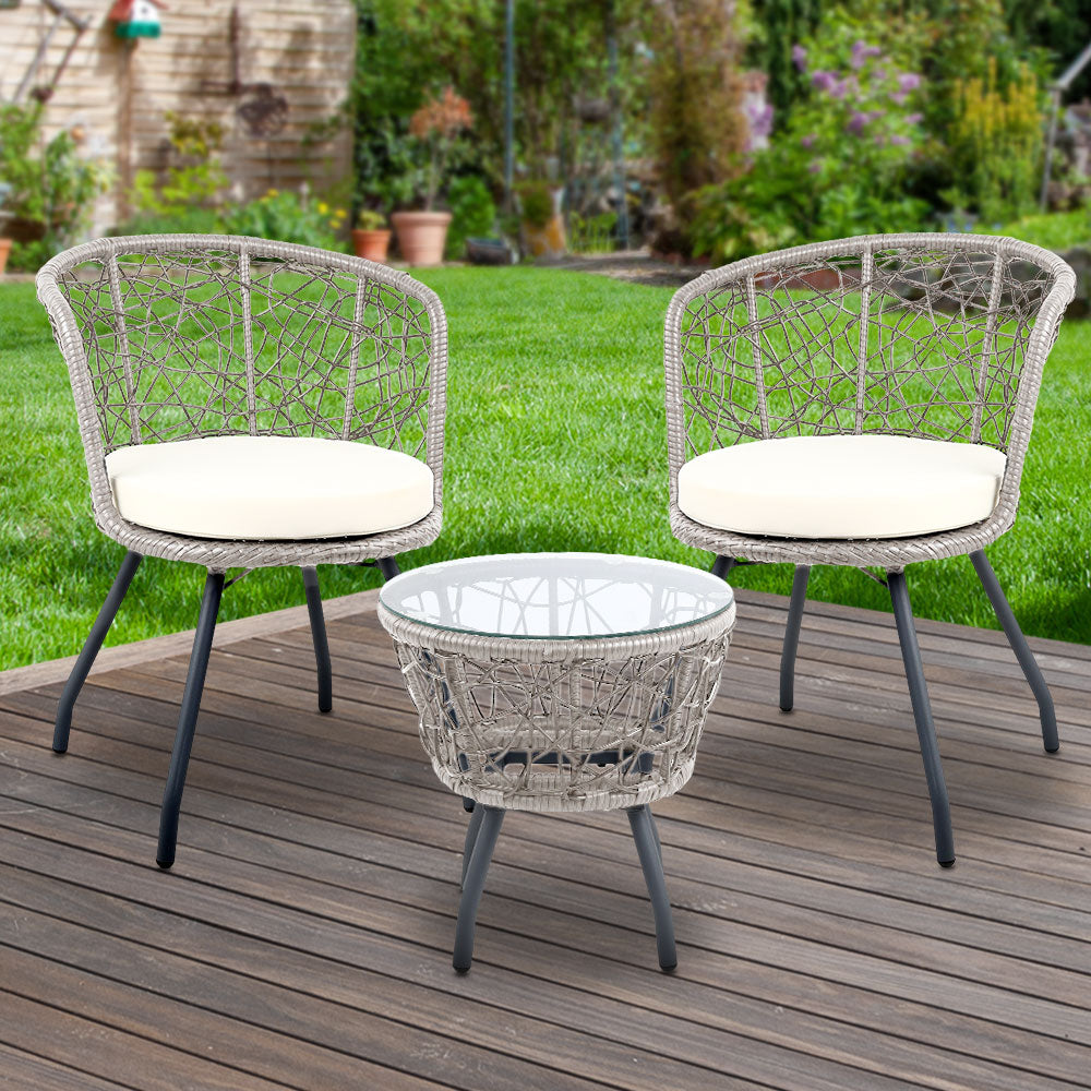 A three-piece outdoor bistro set comprising two high-backed chairs with beige cushions and a round rattan table with a clear glass top on a grey frame, on a wooden deck in a garden setting with green lawn, trees and flowers in the background.