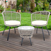 A three-piece outdoor bistro set comprising two high-backed chairs with beige cushions and a round rattan table with a clear glass top on a grey frame, on a wooden deck in a garden setting with green lawn, trees and flowers in the background.